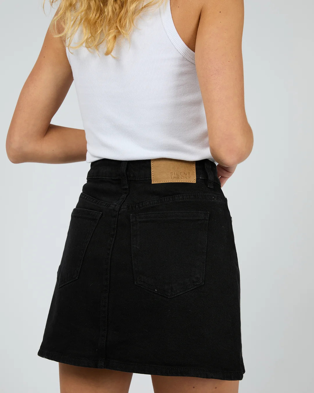 Rear view of a person wearing a black denim mini skirt with a tan leather brand patch at the waistband, paired with a white ribbed tank top, against a light gray studio background.